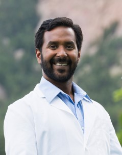Smiling Boulder Dental Center dentist in a white coat, photographed outdoors in Boulder with trees and rock formations behind him